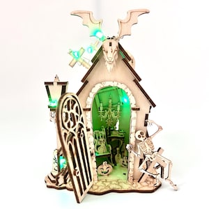 May include: A wooden Halloween-themed house with a bat and skull design on top. The house has an open door, a skeleton, and a pumpkin. The interior is lit with green lights, and a chandelier is visible inside.