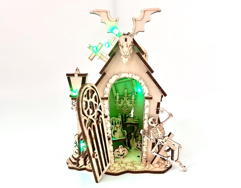 May include: A wooden Halloween-themed house with a bat and skull design on top. The house has an open door, a skeleton, and a pumpkin. The interior is lit with green lights, and a chandelier is visible inside.