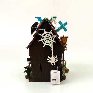 May include: A miniature Halloween-themed house with a dark brown exterior, adorned with a white spiderweb, a hanging spider, and a string of lights. The roof features a black bat, blue crosses, and a decorative lamp post. The house is lit with green lights.