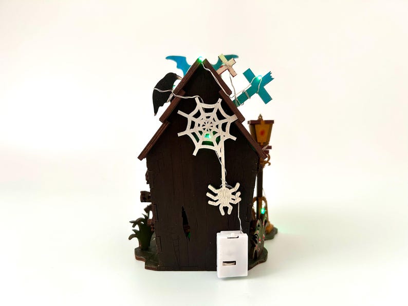May include: A miniature Halloween-themed house with a dark brown exterior, adorned with a white spiderweb, a hanging spider, and a string of lights. The roof features a black bat, blue crosses, and a decorative lamp post. The house is lit with green lights.