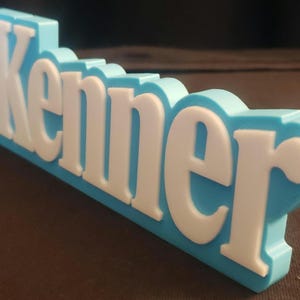 May include: A three-dimensional sign in light blue with the word "Kenner" in white lettering. The sign has a retro design, with the letters slightly raised from the background. The sign is made of a solid material, possibly plastic.