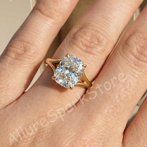 May include: A gold ring featuring a large, rectangular-cut diamond. The ring has a split shank design, with the band splitting into two delicate prongs that cradle the diamond. The diamond sparkles brightly, reflecting light.
