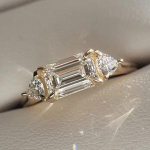 May include: A gold ring with an emerald-cut central gemstone and two pear-shaped side stones. The ring is set in a gold band and presented in a white jewellery box. The gemstones sparkle, reflecting light.