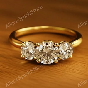 May include: A gold ring featuring three clear, round gemstones. The ring is set on a wooden surface. The text "AllureSparkStore" is visible in the background.