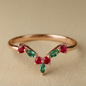 Könnte beinhalten: Ein V-förmiger Goldring mit einer Anordnung aus drei runden roten Edelsteinen und zwei grünen Edelsteinen im Marquise-Schliff in symmetrischem Muster. Die Ringschiene ist schmal und poliert, wobei jeder Stein in einer Krappenfassung sitzt.