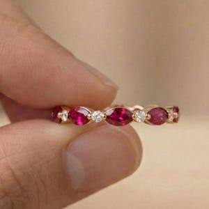 May include: A gold band ring featuring an alternating pattern of marquise-cut deep red rubies and small round brilliant-cut white diamonds. The stones are set in a continuous row along the metal band, creating a delicate and symmetrical design.