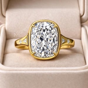May include: A gold ring with a large, rectangular, clear gemstone. The ring has a gold band with small accent stones on each side of the main stone. The ring is displayed in a beige ring box.