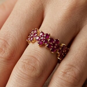 May include: A gold band ring featuring a continuous row of small, round, deep pinkish-red gemstones arranged in a floral pattern. The ring is worn on a finger, showcasing the metallic gold setting and the vibrant, saturated color of the stones.