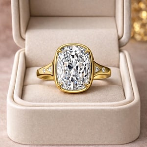 May include: A gold ring featuring a large, cushion-cut clear gemstone set in a bezel setting. The band includes two small triangular accents on each side, each embedded with a tiny round clear stone, creating a symmetrical design.