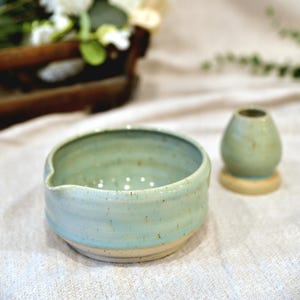 Peut inclure: Un bol à thé en céramique avec un glaçage bleu clair et un petit bec verseur, à côté d'un petit support de fouet à thé assorti. Le bol a une base beige clair. Un plateau en bois avec des fleurs est en arrière-plan.