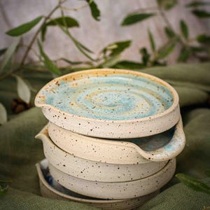 Puede incluir: Una pila de cinco platillos de cerámica con un esmalte blanco moteado y un diseño de remolino azul y verde. Los platillos tienen un pequeño pico de vertido en un lado.