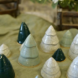 Op de afbeelding: Een verzameling keramische kerstboomfiguren in verschillende tinten groen en wit. De bomen hebben een glanzende afwerking en zijn gerangschikt op een lichtgroen oppervlak. Sommige bomen hebben een gelaagd ontwerp, andere zijn glad.