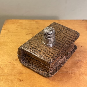 May include: A silver thimble sits atop a small, rectangular, brown leather case. The thimble has a detailed, textured design. The case has a reptile-like pattern and a small clasp. The items are on a light brown wooden surface.