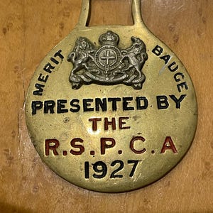 May include: A circular brass horse brass featuring a raised crest with a crown and animals. The text reads MERIT BADGE, PRESENTED BY THE R.S.P.C.A, and 1927. The metal has a polished finish with black and red enamel lettering accents.