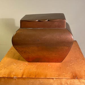 May include: A copper-coloured, antique-style container with a square lid. The container has a rounded body and sits on a wooden surface. The copper has a warm, reddish-brown hue, and the lid fits snugly on top. The container is likely used for storage.
