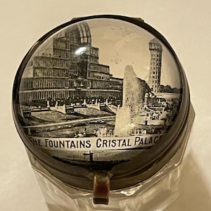 May include: A clear glass inkwell with a hinged metal lid featuring a black and white photograph under a glass dome. The image depicts a large building and a fountain with the text THE FOUNTAINS CRISTAL PALACE printed along the bottom edge.