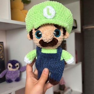 May include: A green crocheted plush toy with a green hat and a white 'L' on the front. The toy has brown yarn eyebrows, a brown yarn mustache, and blue yarn overalls.