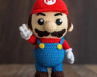 Pode incluir: Um brinquedo de pelúcia de crochê do personagem de videogame Mario. Ele está vestindo um chapéu vermelho com um 'M' branco, uma camisa vermelha, um macacão azul e sapatos marrons. Ele tem um bigode marrom e uma expressão amigável.