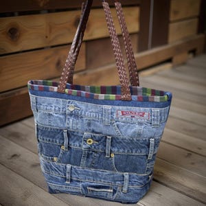 May include: A tote bag constructed from repurposed blue denim jeans, featuring multiple horizontal panels, metal rivets, and a visible Silver brand label. The bag includes two brown braided handles and a colorful striped fabric lining at the top.