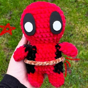 Könnte beinhalten: Handgefertigte, rote Deadpool-Puppe aus Häkelarbeit mit schwarzen Akzenten und großen weißen Augen. Die Puppe hat einen braunen Gürtel und ist etwa 15 cm groß. Ein einzigartiger, handgefertigter Artikel.