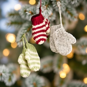 Op de afbeelding: Drie miniatuur gebreide wantenornamenten hangend aan touwtjes. Eén want is rood-wit gestreept, één is effen lichtgrijs met een gespikkelde textuur en één is lichtgroen-wit gestreept. Elke want is voorzien van een klein duimgedeelte.