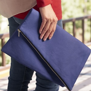 May include: A solid navy blue fold-over clutch bag featuring a silver-toned metal zipper closure that runs diagonally across the front. The rectangular pouch is constructed from a smooth, matte fabric and maintains a clean, simple design.