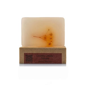May include: A rectangular bar of soap with a light amber hue, featuring a delicate sprig of dried flowers embedded within. The soap rests on a brown cardboard box with text. The soap is a natural skincare product.