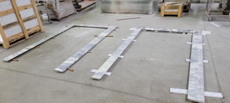 May include: Two rectangular frames made of white marble tiles are laid out on a concrete floor. The frames are made of individual tiles that are arranged in a grid pattern. The frames are positioned at a 90-degree angle to each other.