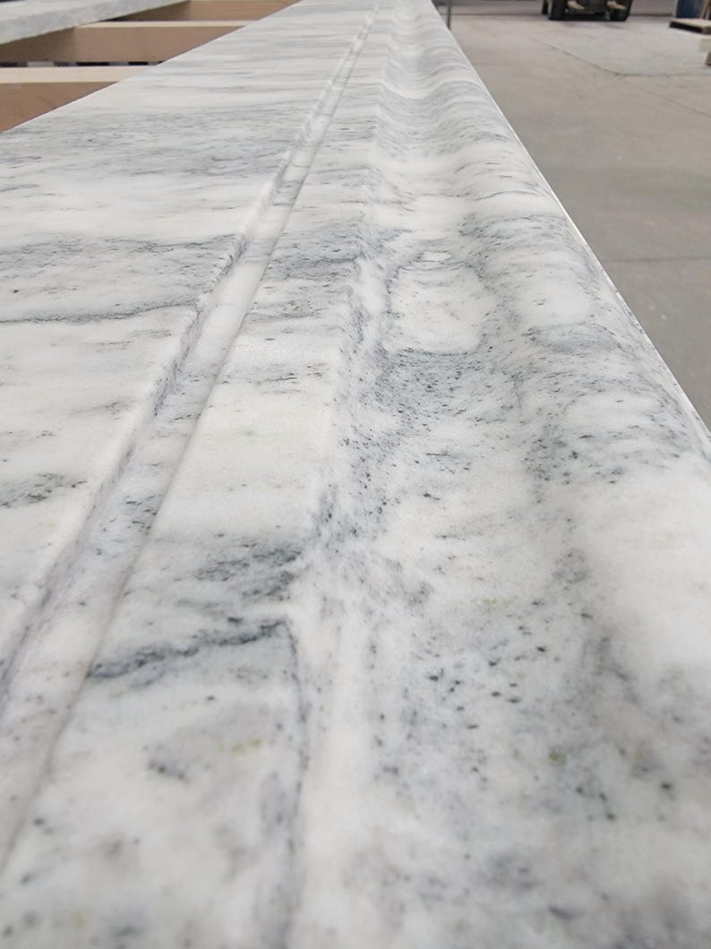 May include: Close-up of a white marble countertop with a decorative edge detail. The marble has a subtle gray veining pattern.