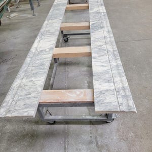 May include: Two large slabs of white marble with gray veining are resting on a metal frame with wooden supports. The marble slabs are on a wheeled cart in a workshop setting.