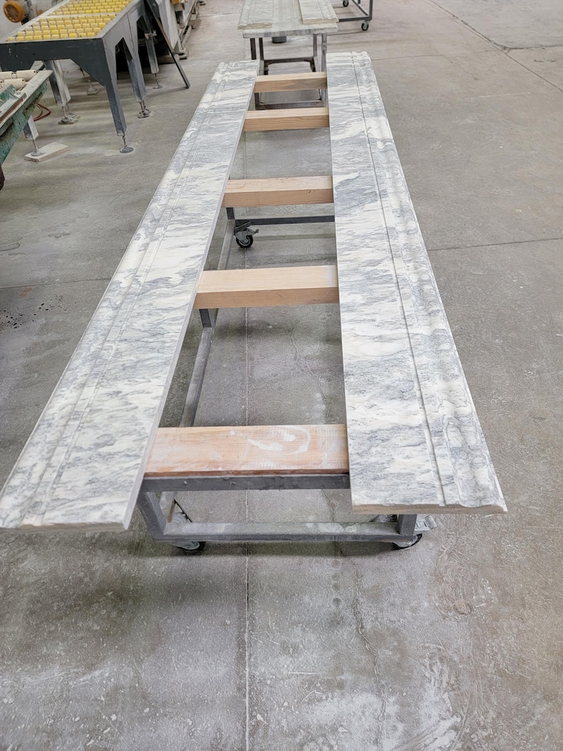 May include: Two large slabs of white marble with gray veining are resting on a metal frame with wooden supports. The marble slabs are on a wheeled cart in a workshop setting.