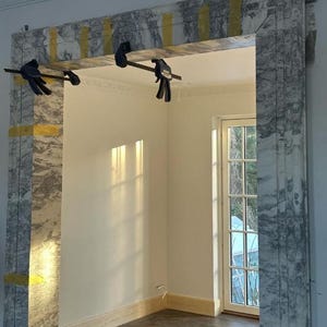 May include: A marble frame is being installed in a room with a white wall and a window. The frame is held in place with clamps. The room has a wooden floor.