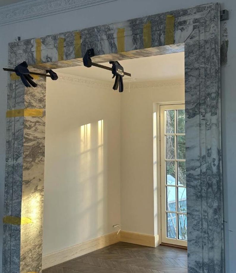 May include: A marble frame is being installed in a room with a white wall and a window. The frame is held in place with clamps. The room has a wooden floor.