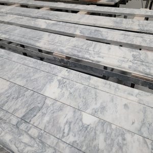 May include: Close-up of a stack of long, rectangular, light gray marble tiles with a white veining pattern. The tiles are stacked on top of each other and are arranged in a staggered pattern.