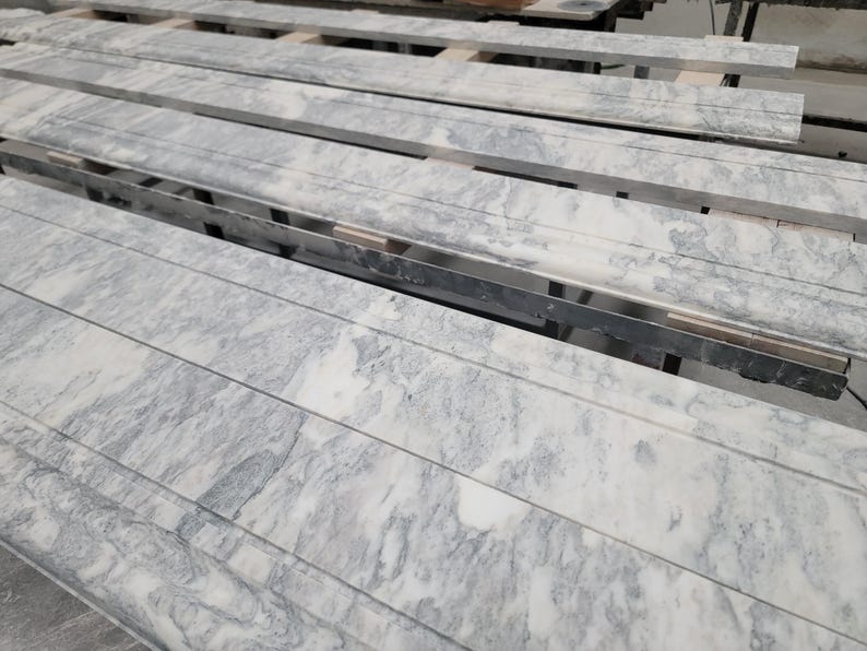 May include: Close-up of a stack of long, rectangular, light gray marble tiles with a white veining pattern. The tiles are stacked on top of each other and are arranged in a staggered pattern.