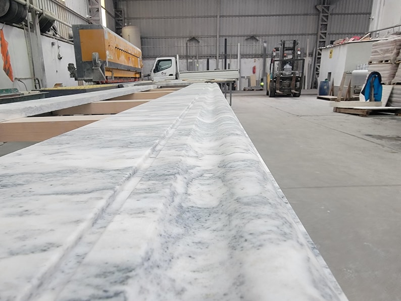 May include: Close-up of a white marble countertop with a decorative edge detail. The countertop is in a workshop setting.