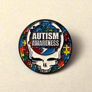 May include: A round pin with a black border featuring a skull design with a white face and teeth. The pin has a colorful jigsaw puzzle border and the words "AUTISM AWARENESS" in white. A blue bear is at the top.