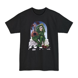 May include: Black t-shirt featuring a seated skeleton in a green jester outfit, holding a ukulele. The illustration is set against an arched backdrop with a checkerboard floor and roses. The design has a vintage, artistic style.