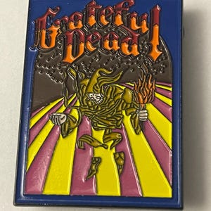 May include: Rectangular pin with a blue border featuring the words "Grateful Dead" in orange and red. The pin's center depicts a skeleton figure holding a torch, set against a yellow and pink striped background.
