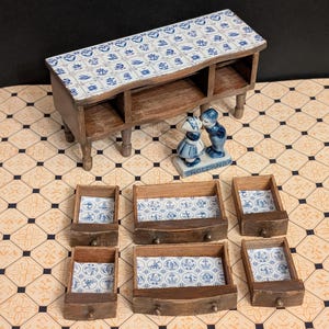May include: Miniature wooden cabinet with six drawers and a Dutch-style blue and white tile top. The drawers and cabinet are brown. A small ceramic figurine of a boy and girl kissing is on the top. The word "HOLLAND" is on the base of the figurine.