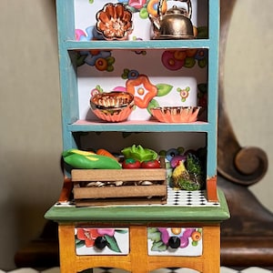 May include: A miniature wooden kitchen hutch with two shelves, painted in a teal blue color. The hutch has a floral patterned wallpaper and features copper-colored kitchenware, including a teapot, bowls, and pots. The hutch sits on a wooden table with two drawers and a lower shelf, painted in a yellow and green color scheme. The table has turned legs and sits on a black and white checkered floor.