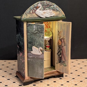 May include: A miniature decorative cabinet with an arched top, featuring a swan and floral details. The cabinet doors open to reveal a rabbit figure and a small interior scene with a table and teacups. The cabinet is painted in various colors.