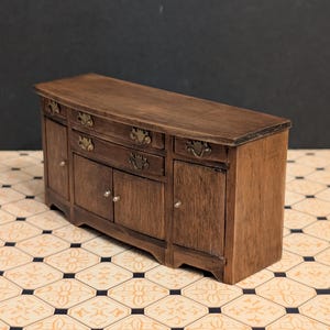 May include: A miniature wooden buffet with a curved top and ornate drawer pulls. The dark brown cabinet has two doors and three drawers. The piece is set on a patterned tile floor.