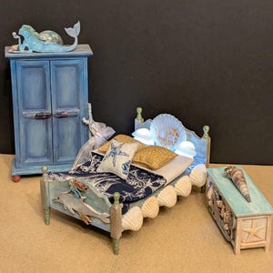 May include: Miniature bedroom set with a coastal theme. Includes a blue painted wardrobe, bed with seashell accents, and a small chest. The bed has a seashell headboard and a starfish pillow. A mermaid figurine sits atop the wardrobe.