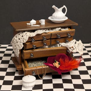 May include: Miniature wooden dresser with drawers, topped with a white pitcher and bowl. A red hat with orange feathers and a gold chain are in the open drawers. A white lace doily is draped over the drawers.