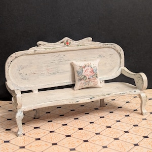 May include: A white, distressed, wooden bench with a floral pillow. The bench has a curved back and ornate details. The pillow has a pink rose and green leaves.