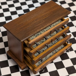 May include: A miniature wooden dresser with four drawers, each partially open. The dresser is stained a dark brown, with silver knobs and a patterned fabric lining the drawers. The piece is set on a black and white checkered surface.