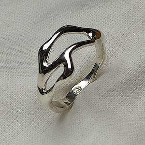 Może przedstawiać: Srebrny pierścionek o abstrakcyjnym, organicznym wzorze. Pierścionek wykonany jest z gładkiego, polerowanego metalu i ma unikalny, płynny kształt.