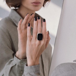 Könnte beinhalten: Eine Frauenhand trägt einen großen, quadratischen Ring aus schwarzem Onyx mit einer silbernen Fassung. Der Ring sitzt am Mittelfinger der Hand.