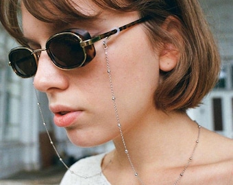 May include: A woman wearing tortoiseshell sunglasses with a silver chain attached to the frames. The chain has small silver beads.
